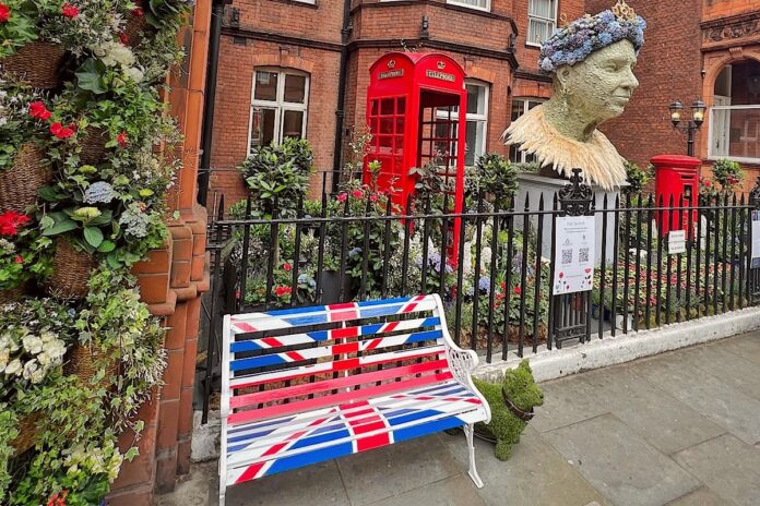 Angielski ogródek w którym znajduje się budka telefoniczna, skrzynka pocztowa, popiersie królowej Elżbiety i ławka pomalowana w brytyjską flagę